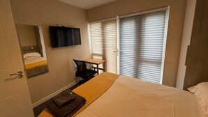A hotel room with a bed, a desk, a chair, a television, and a window with blinds. - Cosy 2-bedroom Flat close to Royal Holloway (London)