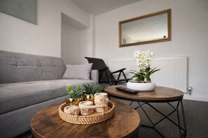 Un salon avec un canapé gris, une table basse ronde en bois agrémentée d’objets décoratifs, une plante en pot et une télécommande.