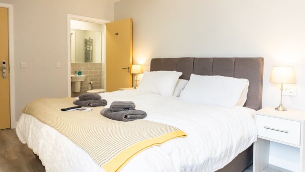 A neatly made bed with pillows and folded towels, a bedside table with a lamp, and a bathroom visible through an open door. - Chic Studio in Cheltenham (Cheltenham)