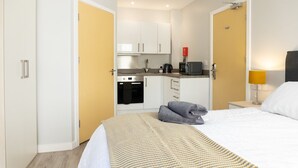 Una habitación de hotel compacta con cama, mesita de noche, lámpara, kitchenette equipada con estufa y microondas, y armario.