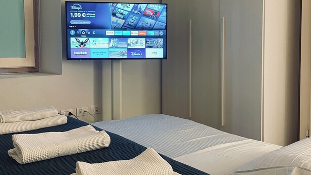 A hotel room with a bed, a TV mounted on the wall displaying various channels, and neatly folded towels on the bed. - The Master's Suite [In the heart of the Borgo] (Bienno)