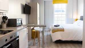 Habitación de hotel compacta con kitchenette, una cama, un televisor y una ventana con persiana amarilla.