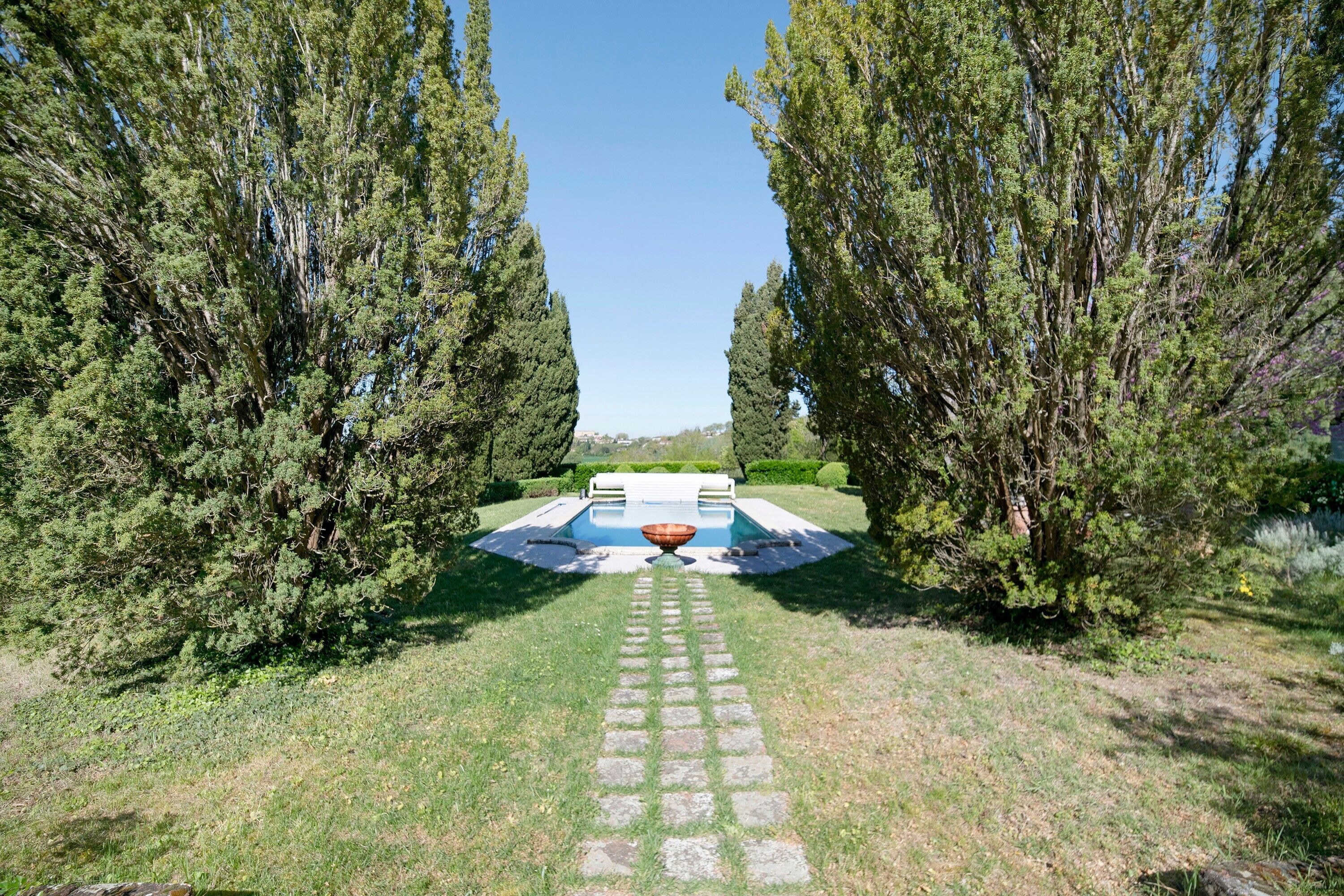 Un sentier mène à une piscine agrémentée d’une fontaine centrale, le tout étant entouré de grands arbres.