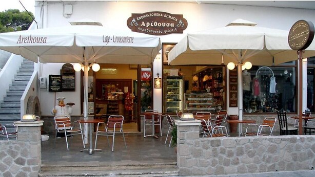 An outdoor café with stone walls, tables, and chairs under umbrellas. - ARETHOUSA (Milos)