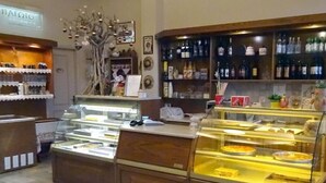 A bakery counter with display cases, a selection of bottles on shelves, and a decorative tree. - ARETHOUSA (Milos)