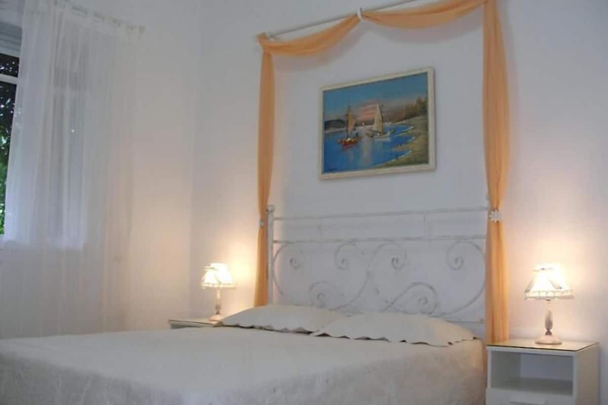 A bedroom with a bed, two bedside tables with lamps, a painting on the wall, and a window with curtains.