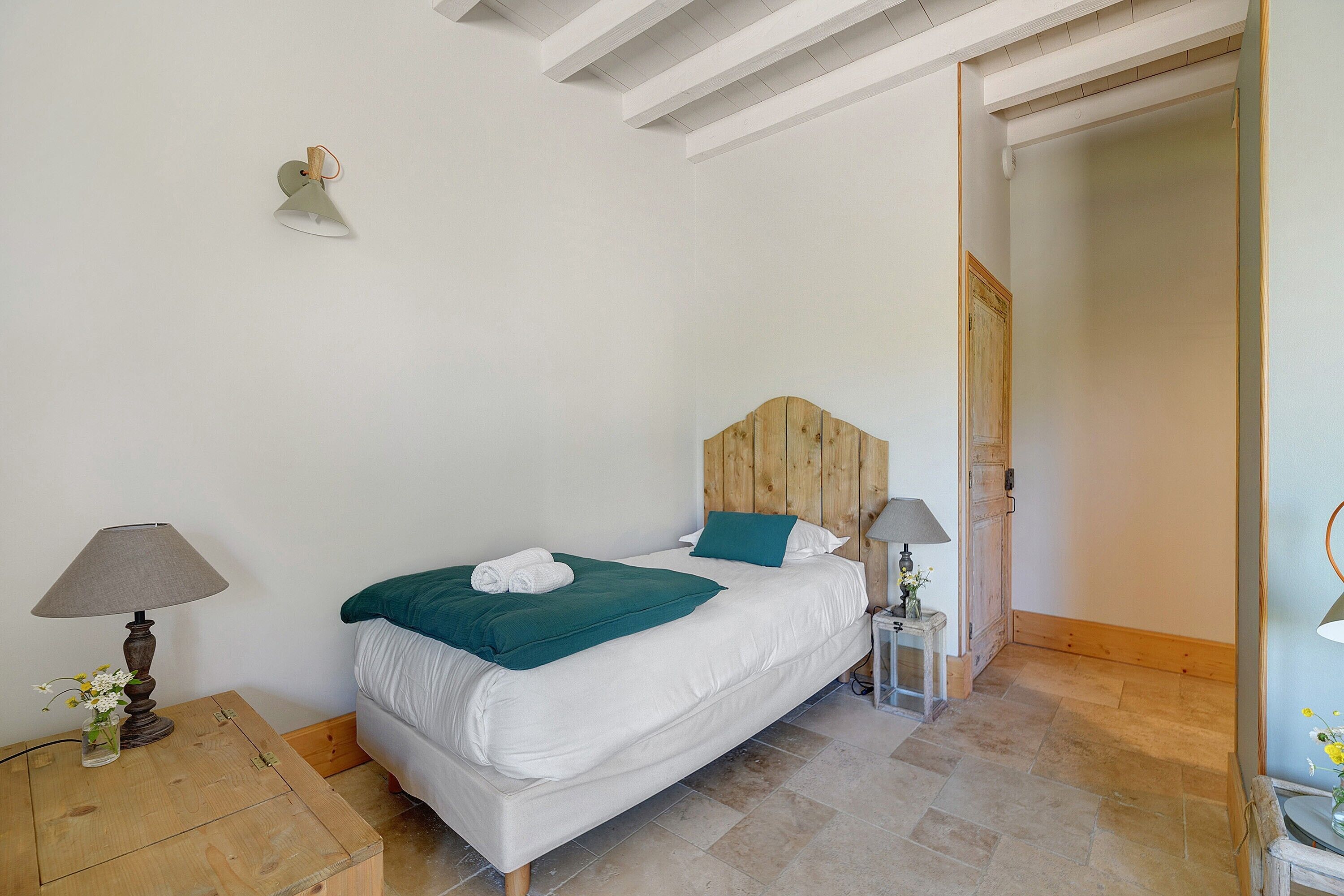 Une chambre à coucher avec une tête de lit en bois, un lit recouvert d’une literie blanche, un plaid vert, deux lampes de chevet et une petite table sur laquelle se trouvent une lampe et des fleurs.