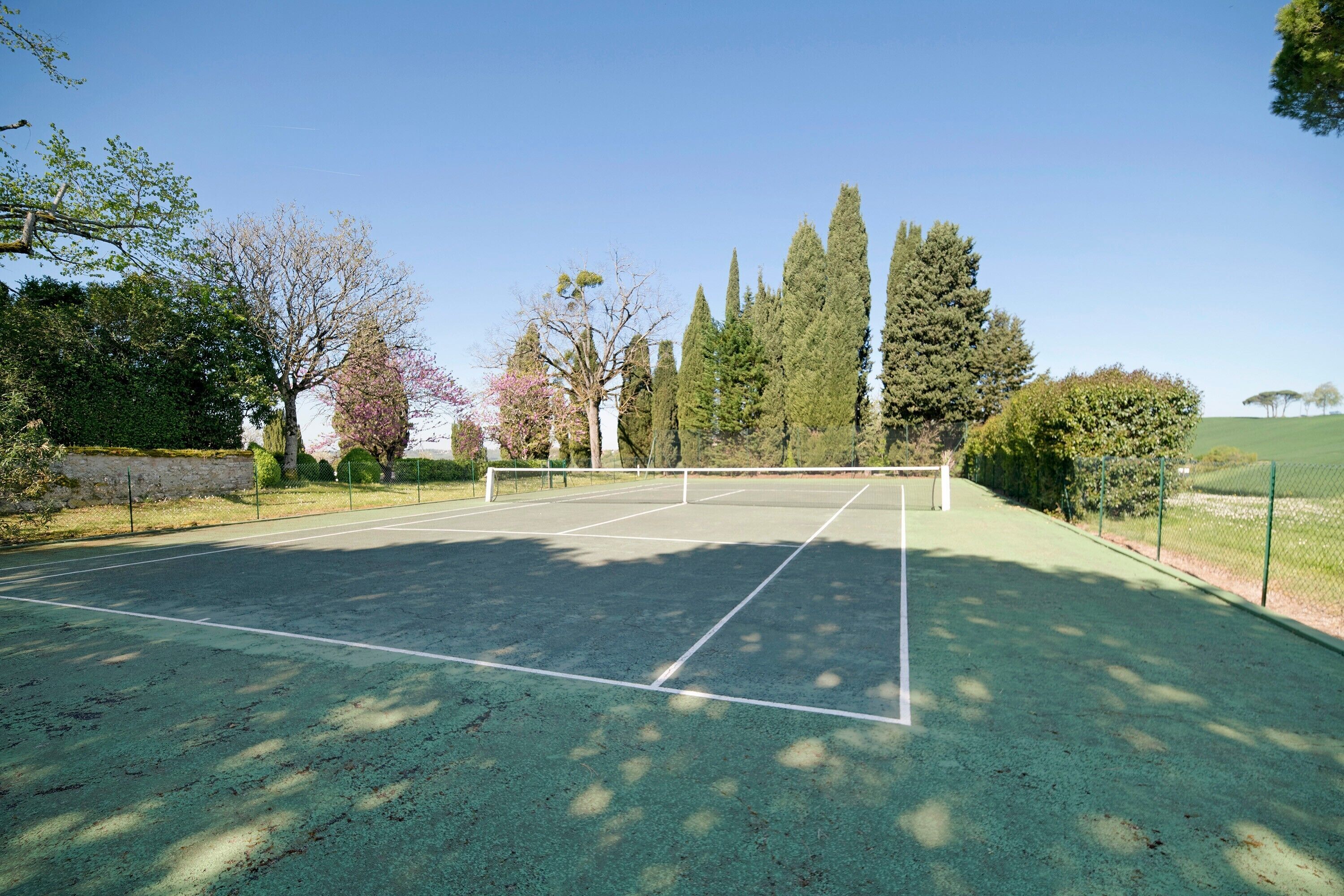 En udendørs tennisbane med net, omgivet af træer og en stenvæg.
