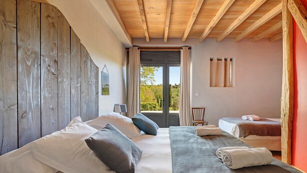 A bedroom with a wooden headboard, a large bed with pillows, a bench, a window with curtains, and a view of greenery outside. - Bed and Breakfast 'Chambre 'Sophie'' with Shared Pool, Wi-Fi and Air Conditioning (Verfeil)