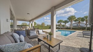 Ein überdachter Außenbereich mit Pool, ausgestattet mit Sofas und einem Tisch, mit Blick auf Palmen und Häuser im Hintergrund.