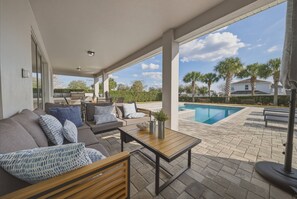 A covered outdoor seating area with a pool, furnished with sofas and a table, and a view of palm trees and houses in the background. - 8BR house sleeps 22 with pool table (Kissimmee)