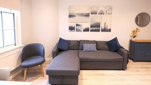 Un salon moderne avec un canapé d’angle gris, un fauteuil, un plancher en bois et un mur orné de tableaux représentant des paysages.