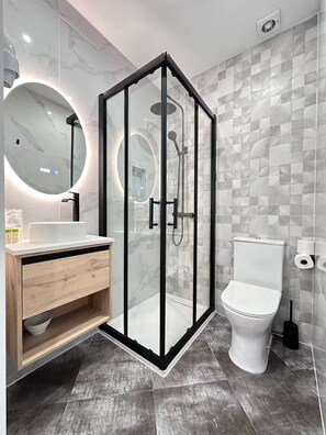 A modern bathroom with a glass-enclosed shower, a round mirror, a white toilet, and a wooden vanity. - Modern Central Ramblas apt: 4 beds & premium baths (Barcelona)