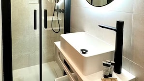Un bagno moderno con cabina doccia in vetro, un lavandino bianco e un rubinetto nero, e uno specchio tondo.