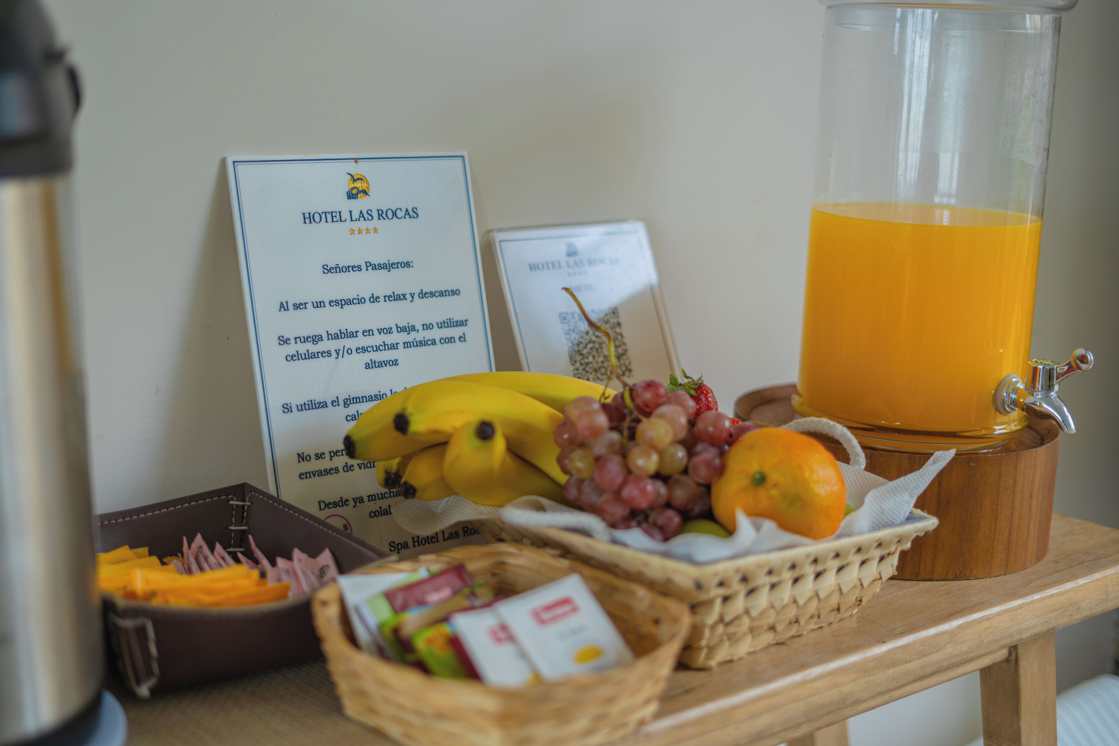 A table with a fruit basket, a glass of orange juice, a sign with hotel information, and a leather bag.
