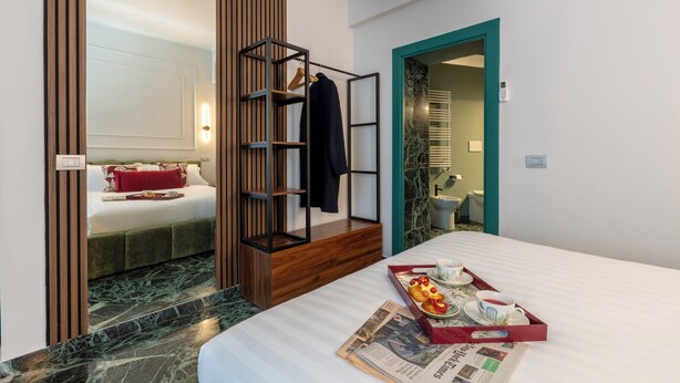 Une chambre d’hôtel avec un lit, une armoire en bois, une étagère avec des vêtements, un plateau avec le petit-déjeuner, et une salle de bain visible à travers une porte ouverte.
