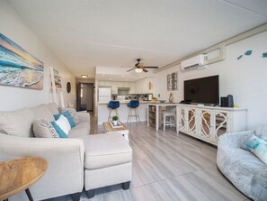 Ein modernes Wohnzimmer mit Sofa, Hocker und Couchtisch. Es gibt eine Küchenzeile mit Esstisch und Stühlen. Ein Deckenventilator und eine Klimaanlage sind zu sehen.