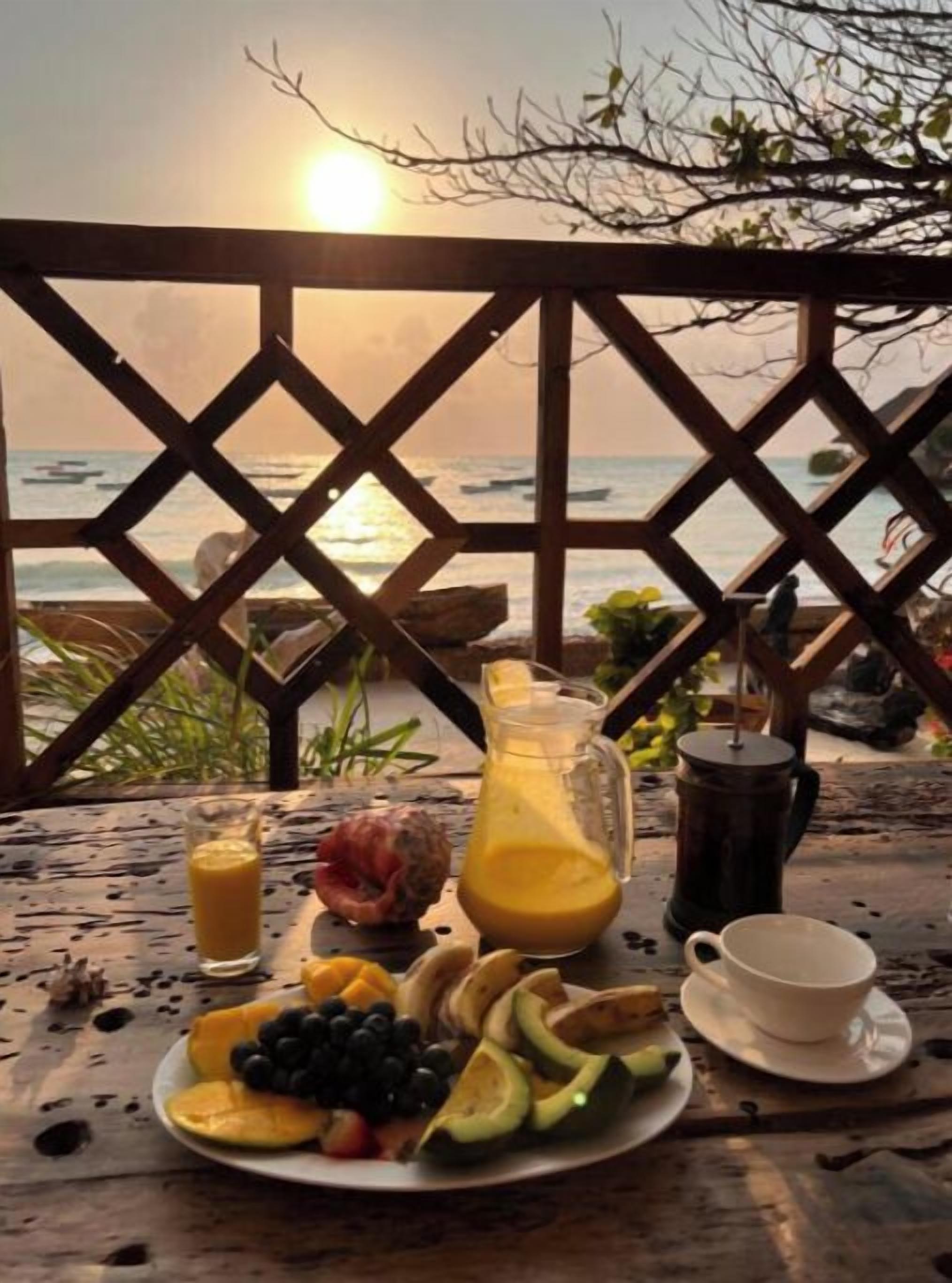 A breakfast table with fruit, a pitcher of orange juice, and a cup, overlooking the ocean at sunset.