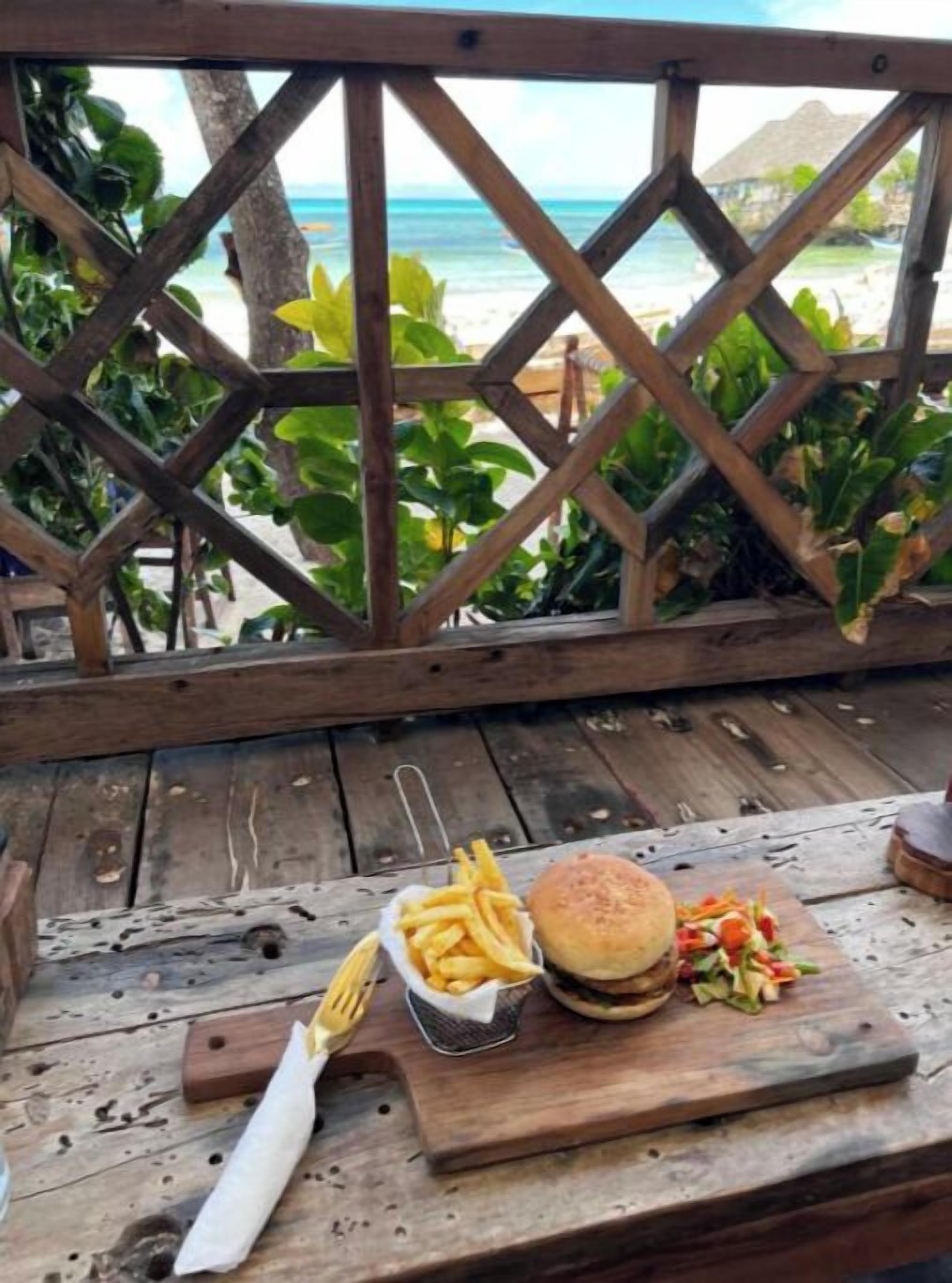 A wooden table with a burger, fries, and salad, set against a wooden fence overlooking a beach.