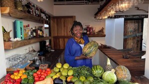Una persona sostiene un melón en un puesto de mercado con diversos vegetales y frutas a la vista.