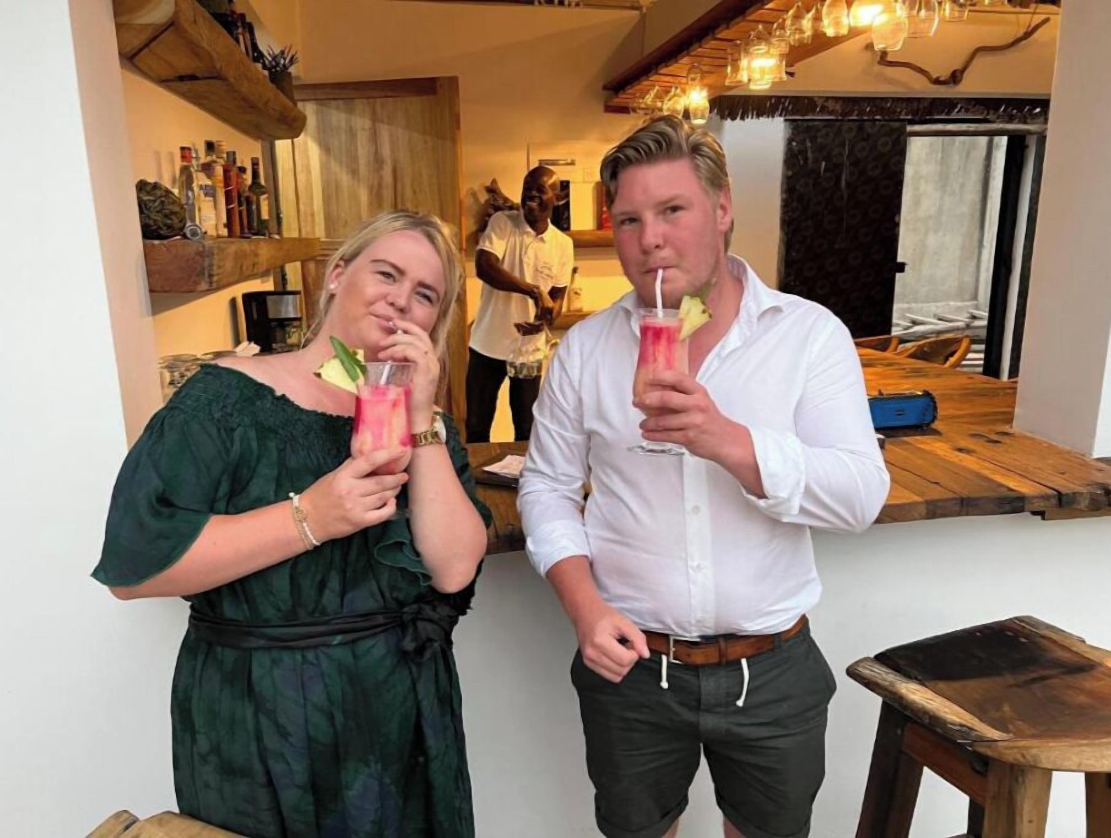 Två personer som håller i drinkar i ett rum med trämöbler och en bardisk.