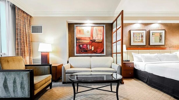 Habitación de hotel con cama, sofá, mesa redonda, una silla, lámpara y obras de arte en las paredes.