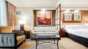 A hotel room with a bed, a sofa, a round table, a chair, a lamp, and artwork on the walls.