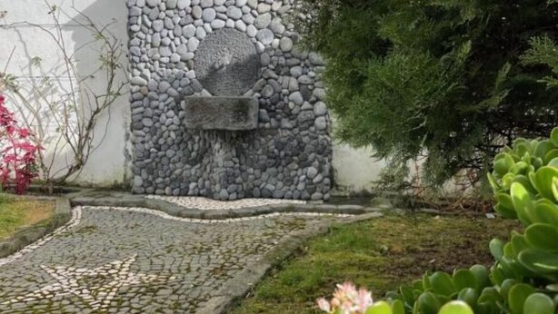Un muro in pietra con un'apertura circolare, un sentiero in pietra e piante verdi.