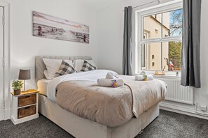 A neatly made bed with a white and beige duvet, a bedside table with a lamp and a small plant, a window with curtains, and a painting on the wall.