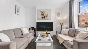 A modern living room with two grey sofas, a white coffee table with breakfast items, a flat-screen TV, and artwork on the walls. - 6 Bedroom House in Warrington Centre Sleeps 12 (Warrington)