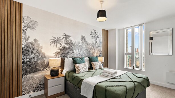 Une chambre à coucher comprenant un lit, une table de chevet, une lampe et une fresque représentant une végétation tropicale sur le mur.