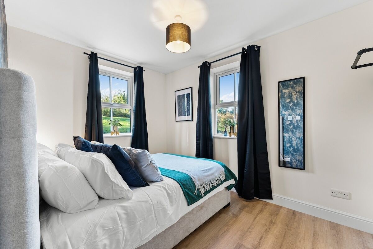 Une chambre à coucher avec un lit, deux fenêtres avec des rideaux, un luminaire au plafond et des œuvres d’art accrochées aux murs.