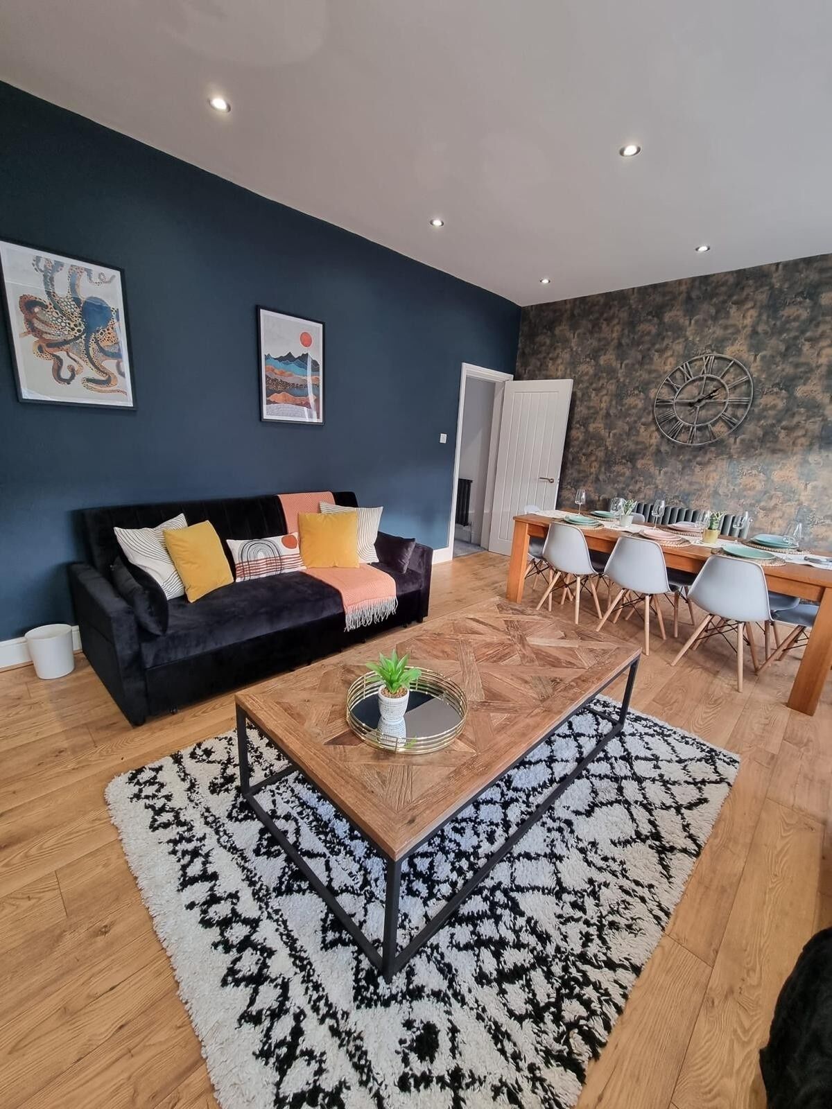 Ein modernes Wohnzimmer mit einer schwarzen Couch, einem Couchtisch mit Pflanze und einem Essbereich mit Holztisch und Stühlen.