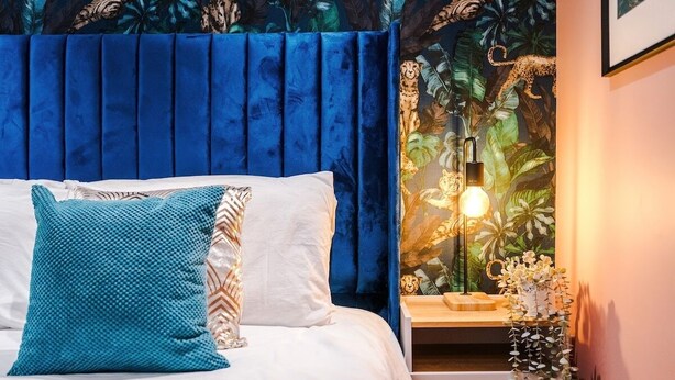 Een slaapkamer met een blauw hoofdeinde, wit beddengoed en een nachtkastje met een lamp en een bloevas.