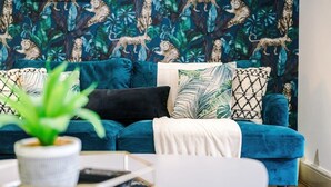Een smaragdgroene bank met gestreepte kussens, een kleine potplant en een witte bijzettafel met een boek en een smartphone.