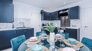 Een moderne keuken met donkere kastjes, een marmeren achterwand en een eethoek met een glazen tafel en blauwe stoelen.