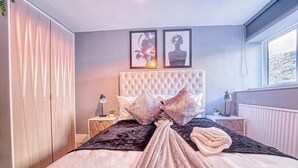 Ein Schlafzimmer mit einem gebuschten Kopfteil, einem Bett mit einem gemusterten Bettbezug, zwei Kissen, einem Nachttisch mit Lampe und einem Fenster mit Blick auf eine verschneite Landschaft.