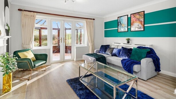 A modern living room with a glass coffee table, a green armchair, a white sofa with blue and green throw pillows, and a teal accent wall. - Large 6 Bedroom Cambridge Abode (Cambridge)