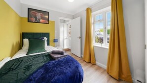 A bedroom with a bed, a green headboard, a framed picture on the wall, and a window with yellow curtains. - Large 6 Bedroom Cambridge Abode (Cambridge)