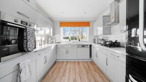A modern kitchen with white cabinets, a built-in oven, a dishwasher, and a large window with an orange blind. - Large 6 Bedroom Cambridge Abode (Cambridge)