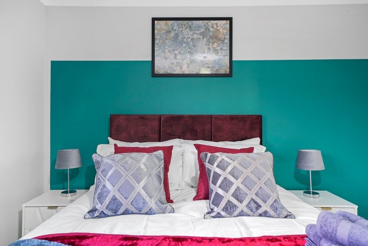 A bedroom with a teal accent wall, a white bed with red and grey patterned pillows, and a framed abstract painting above the bed.