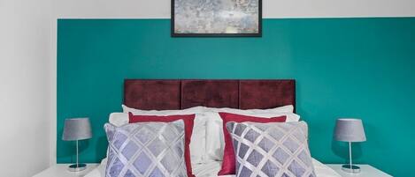 A bedroom with a teal accent wall, a white bed with red and grey patterned pillows, and a framed abstract painting above the bed.