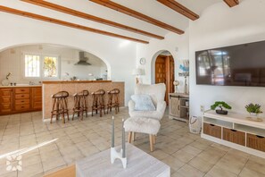 A spacious living area with a large TV, a cozy armchair, and a kitchen with wooden cabinets and a brick island. - Villa SOL Y MAR by Abahana Villas (Benissa)