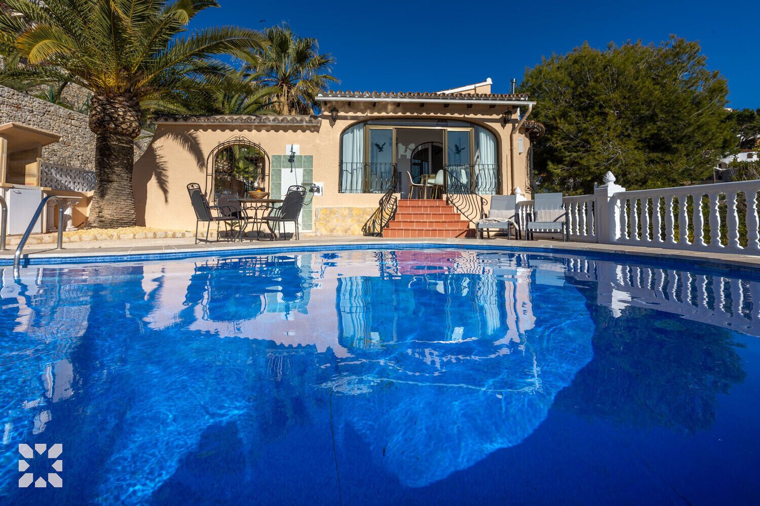A pool area with a clear blue pool, outdoor seating, and a building with a tiled roof and stone walls.