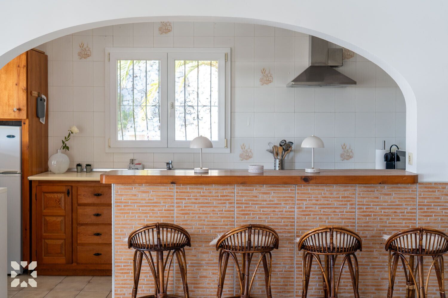 Une cuisine avec un mur recouvert de briques, des meubles en bois, un évier à deux bacs et trois chaises en osier.