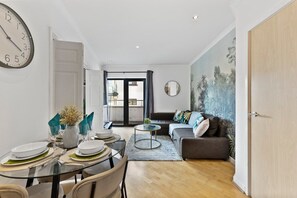 A modern living room with a sofa, a dining table set for four, a round coffee table, and a large wall clock.