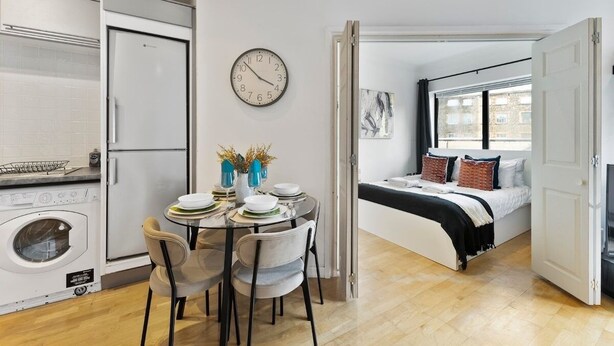 Een compact appartement met een eethoek, een bed, een wasmachine en een groot raam.