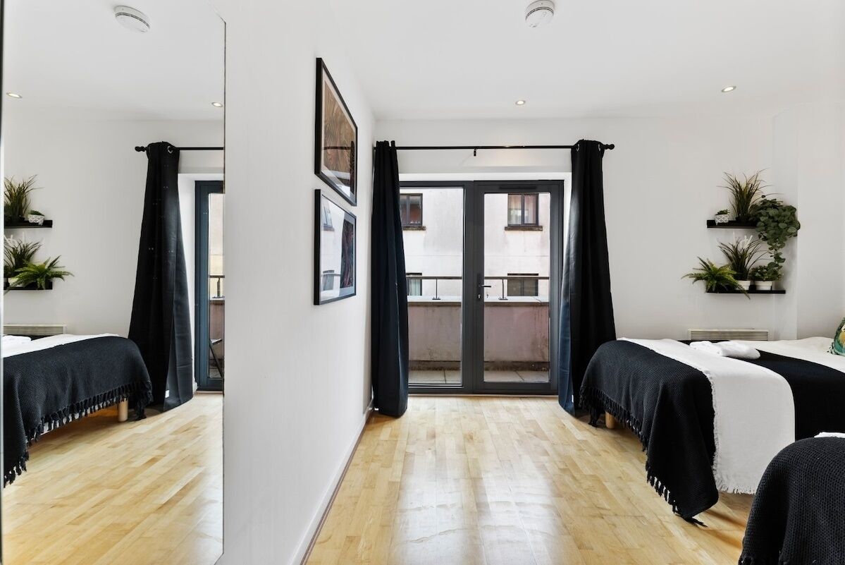 A hotel room with a wooden floor, two beds with black and white bedding, a balcony with a view, and potted plants on the walls.