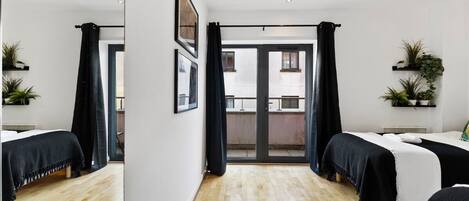 A hotel room with a wooden floor, two beds with black and white bedding, a balcony with a view, and potted plants on the walls.