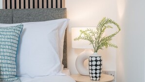 A neatly made bed with white and blue pillows, a round side table with a black and white polka dot vase, and a wall-mounted lamp. - Comfort & Style in Salford - Sleeps 8 (Salford)